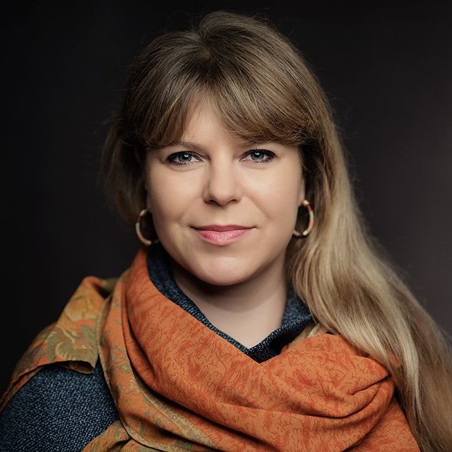 zdjęcie przedstawia Aleksandrę Zińczuk, redaktorkę naczelną magazynu Kultura Enter. To sympatyczna młoda kobietą z długimi blond włosami. Ma kolczyki. Ubrana jest w szarą bluzkę i pomarańczowy szalik