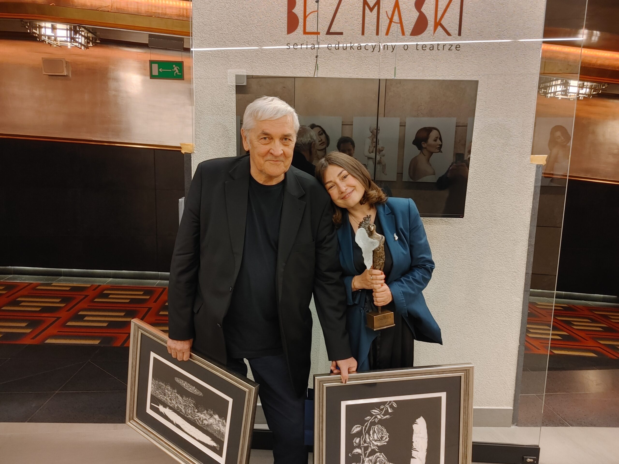 Kobieta i mężczyzna na wystawie "Bez Maski. Seria edukacyjna o teatrze". Mężczyzna trzyma w rękach dwa obrazy, a kobieta statuetkę.
