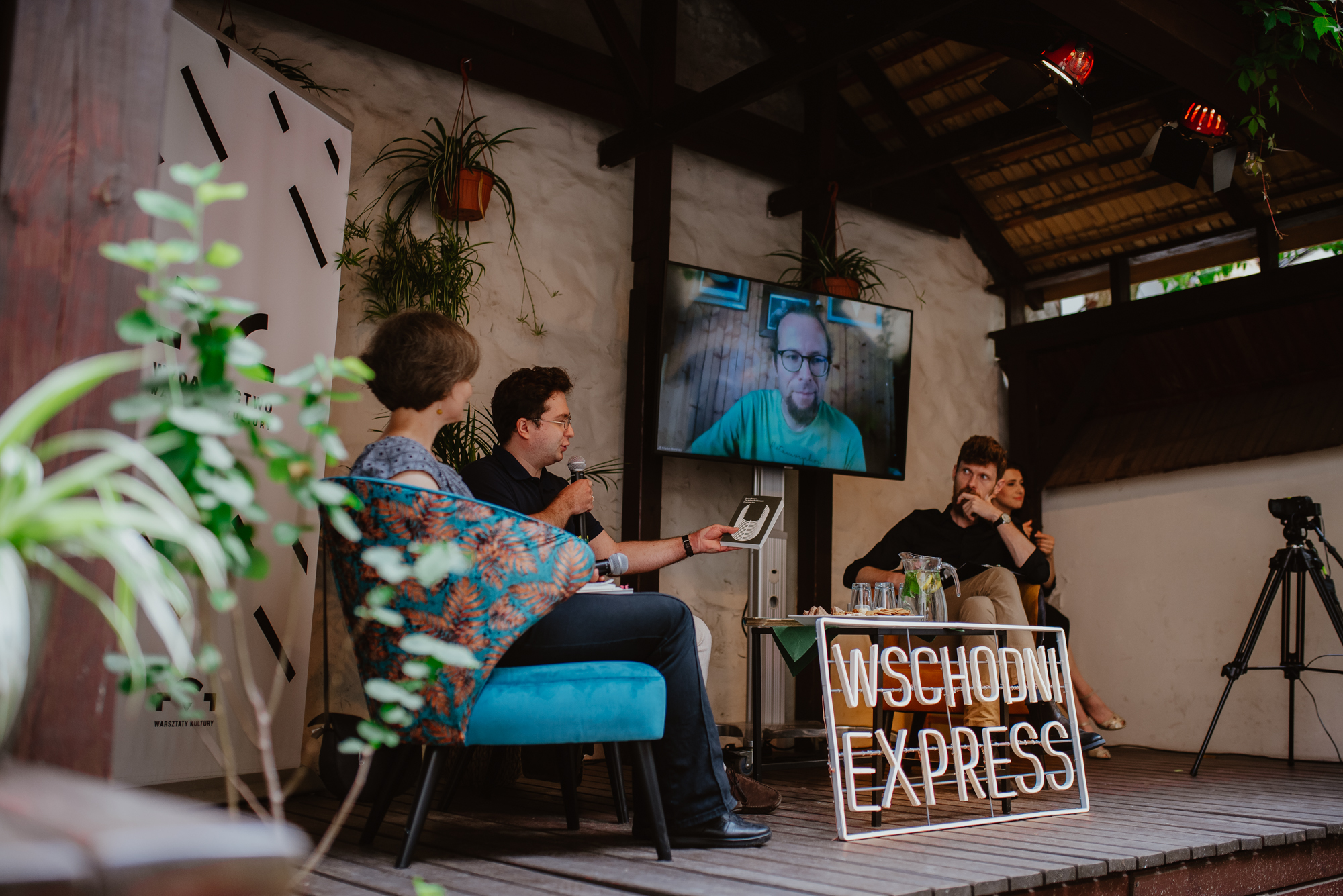 Spotkanie literackie z Mariusem Burokasem. Autor jest wyświetlony na telewizorze. Na scenie tłumaczka Agnieszka Rembiałkowska, moderator Rafał Rutkowski oraz lektor Jan Tuźnik. Przed goścmi ledon Wschodniego Expressu,