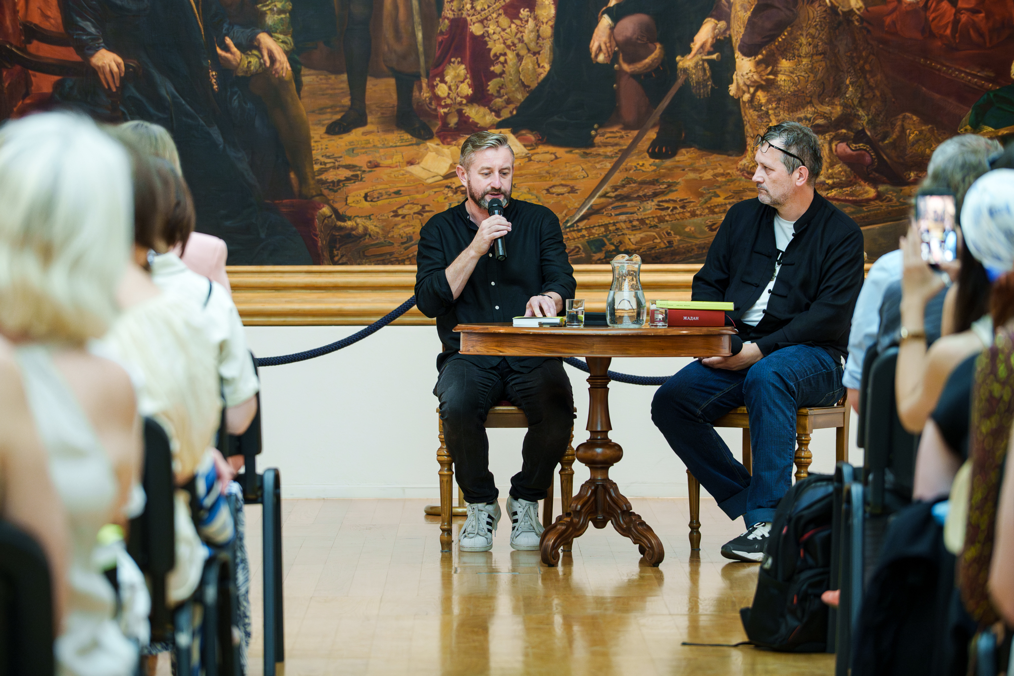 fot. Marcin Butryn (38) Autor książki i inny mężczyzna siedzą przy stole i rozmawiają. W tle jest fragment obrazu.